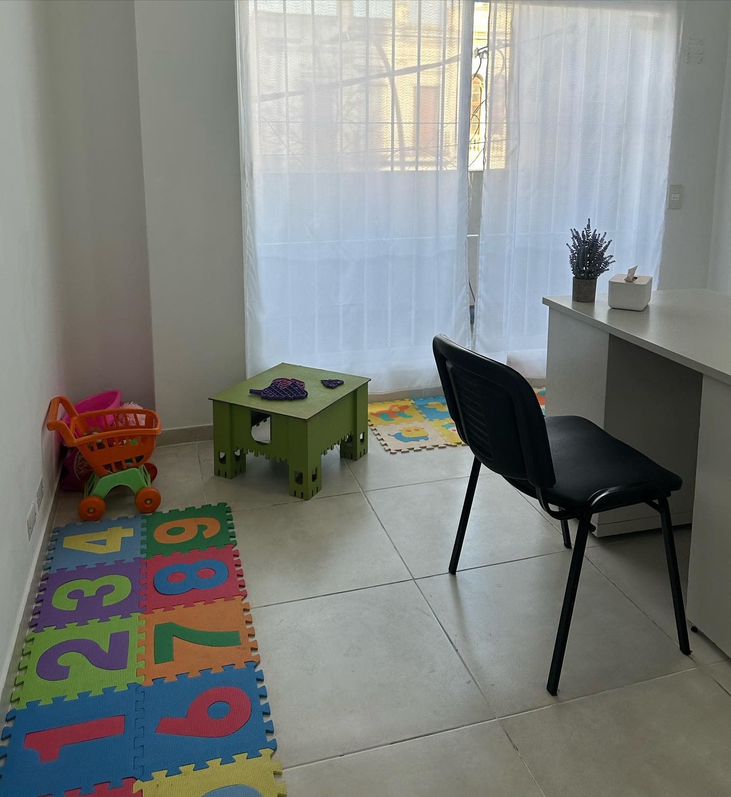Consultorio de Samary con rincón de juego infantil: alfombra de números, mesita verde y escritorio junto a la ventana